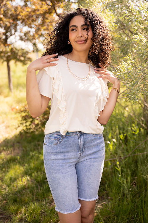 Woman wearing a white blouse and frayed bermuda shorts in front of trees
